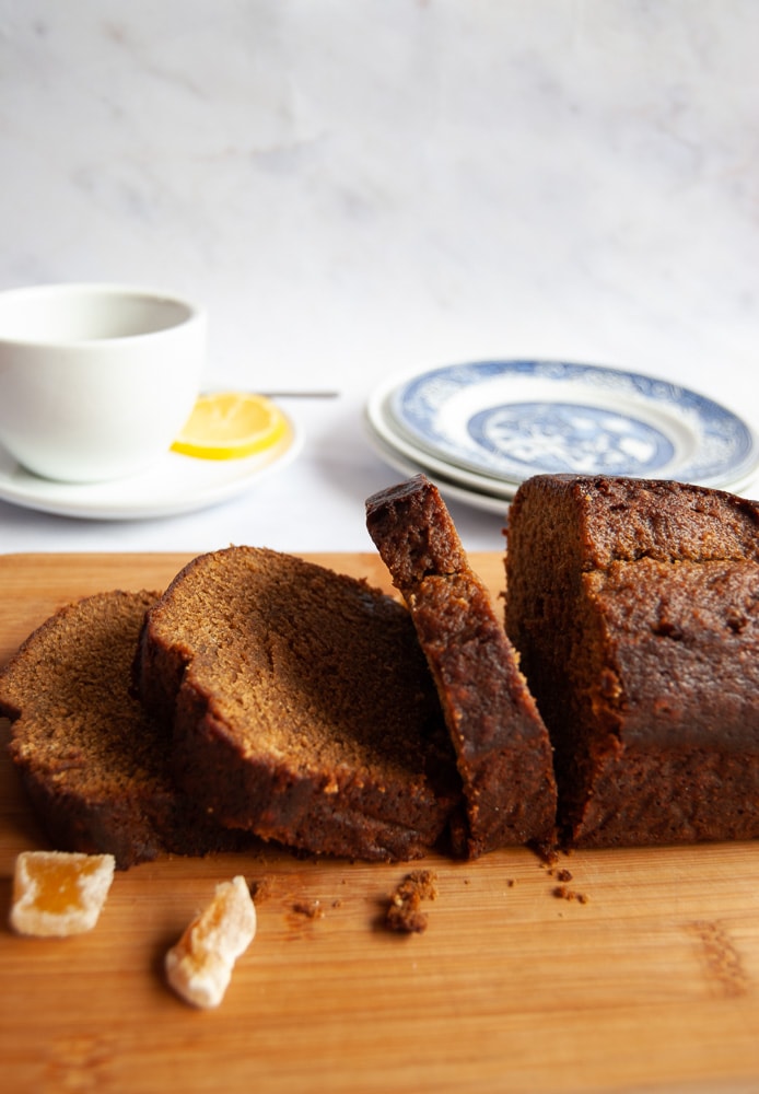 A partially sliced gingerbread loaf on a wooden board. A white cup and saucer of tea and blue Willow pattern plates can be seen in the background.