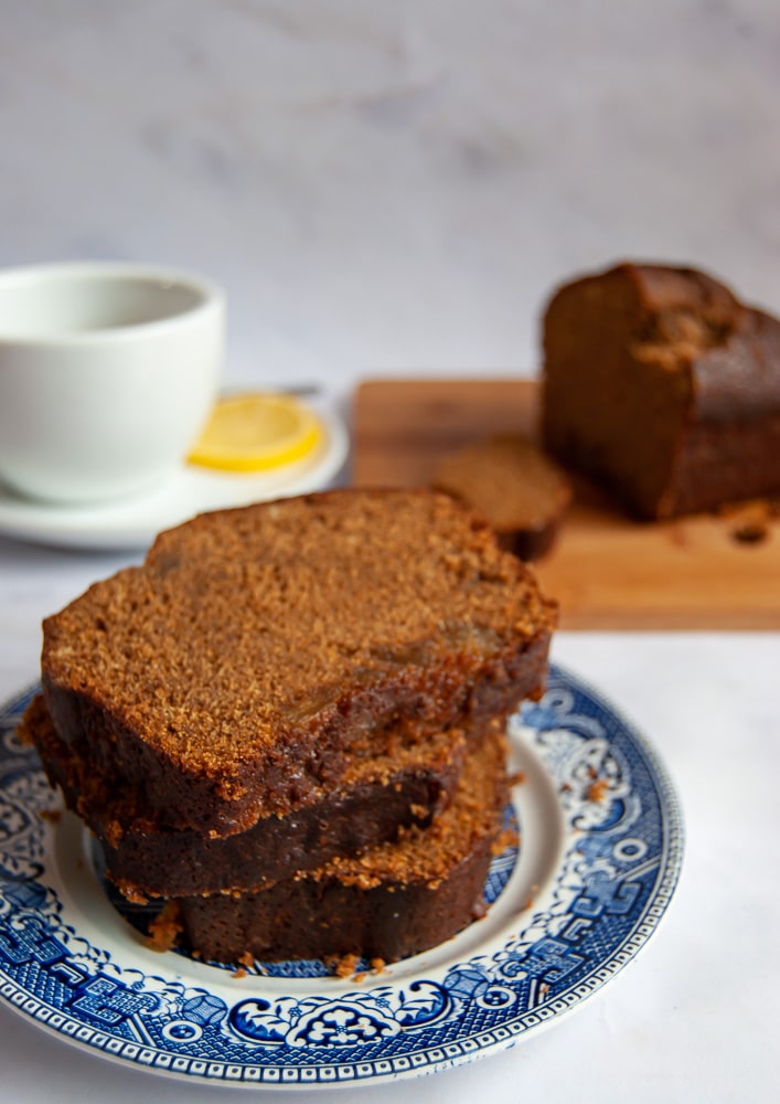 Slices of gingerbread on a blue willow pattern plate/white background. A cup of tea and the rest of the gingerbread can be seen in the background.