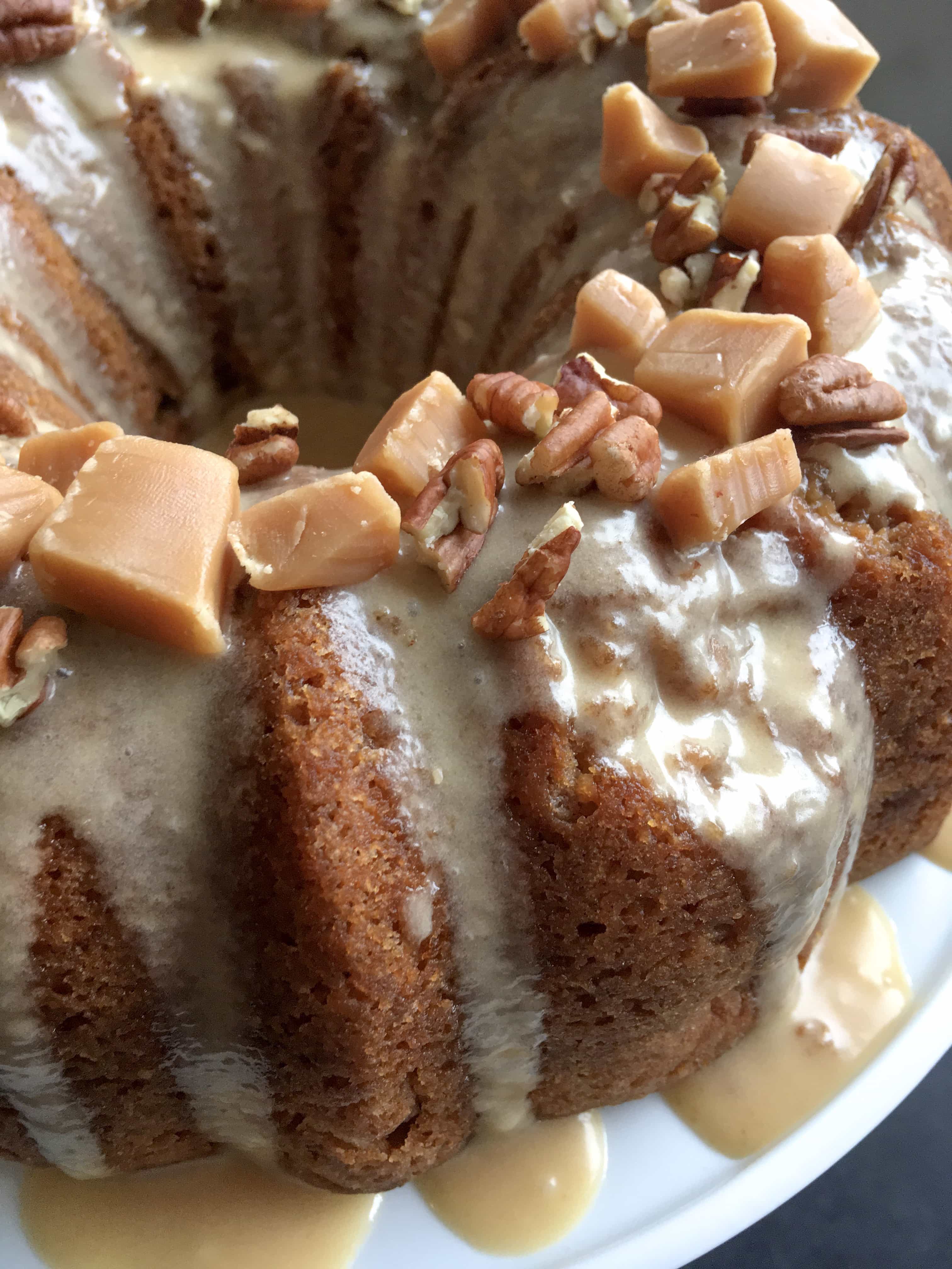 A close up photo of a double apple caramel bundt cake covered in a sticky caramel glaze and topped with fudge chunks and chopped pecans