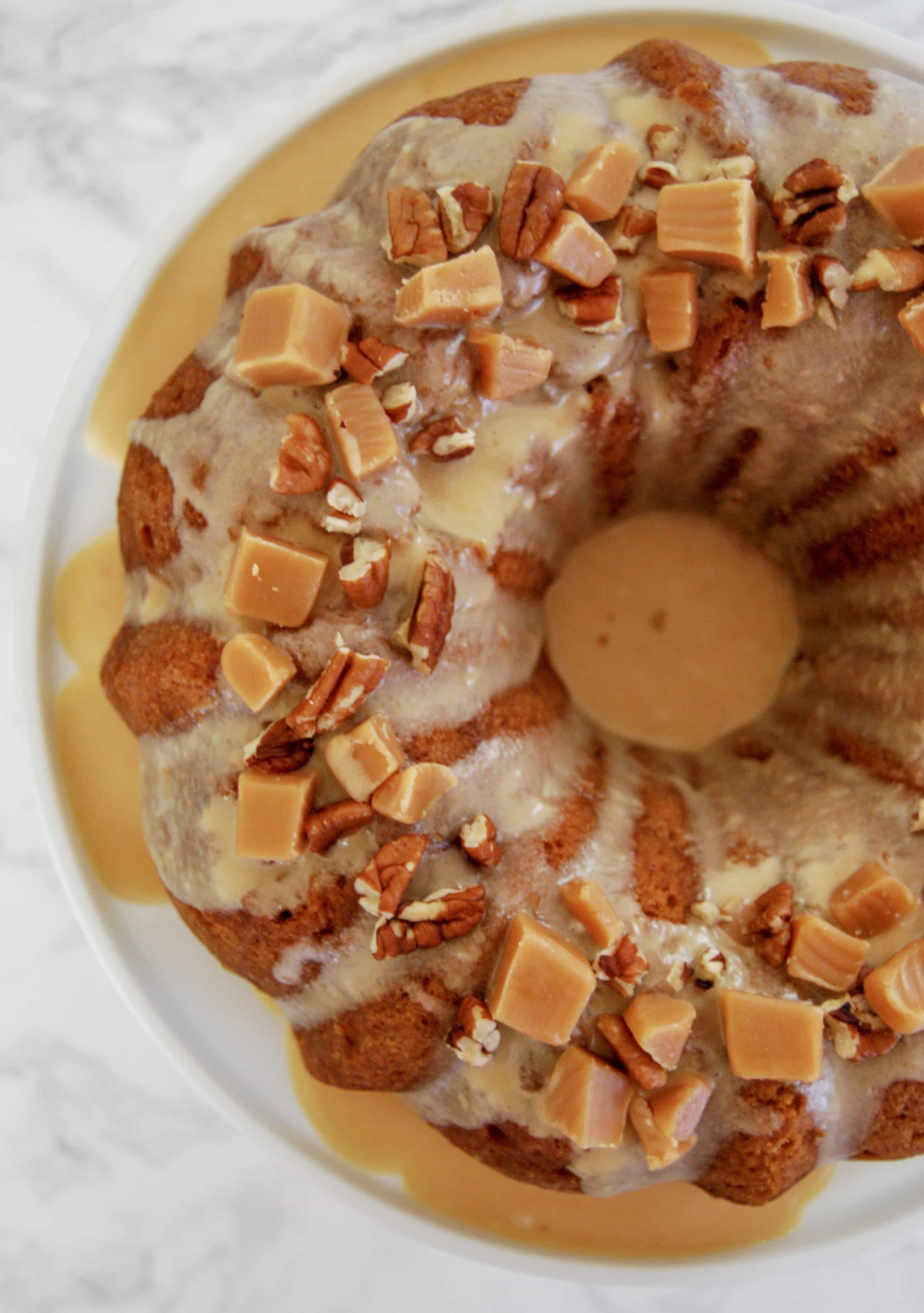 A flat lay photo of an apple caramel bundt cake topped with caramel glaze and topped with fudge pices and chopped pecans