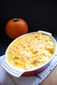 A dish of macaroni cheese with roasted butternut squash. The macaroni is golden and bubbling. A pumpkin can be seen in the background of the photo.