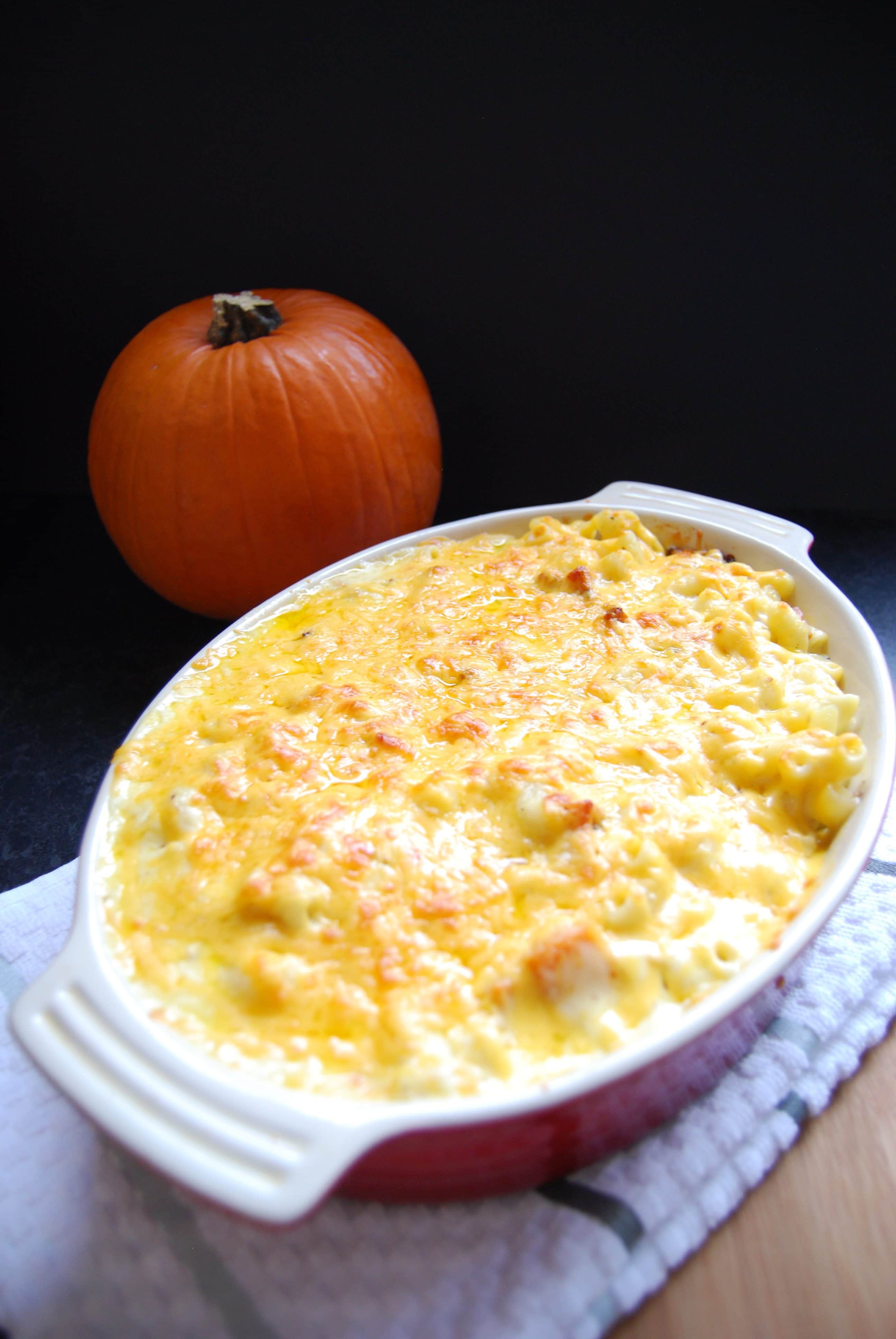 A dish of roasted pumpkin macaroni cheese. The macaroni is golden and bubbling. A pumpkin can be seen in the background of the photo.