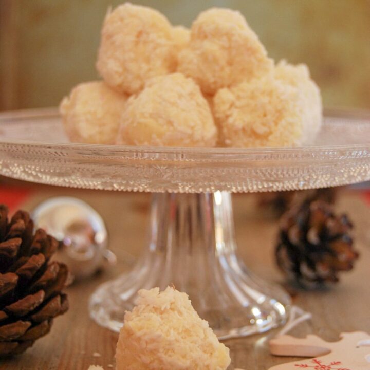 A glass cake stand with a pile of white chocolate coconut truffles on top. Christmas pine cones and red and white Christmas decoration reindeers in the foreground.