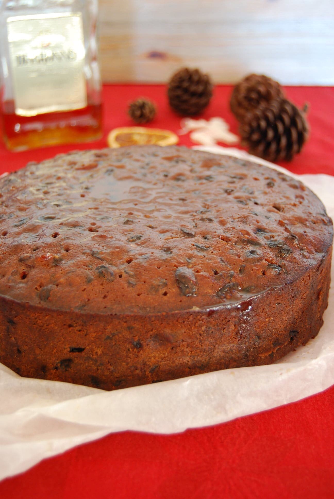A fruit cake partially wrapped in baking parchment. A bottle of Amaretto and pine cones/orange slices can be seen in the background.