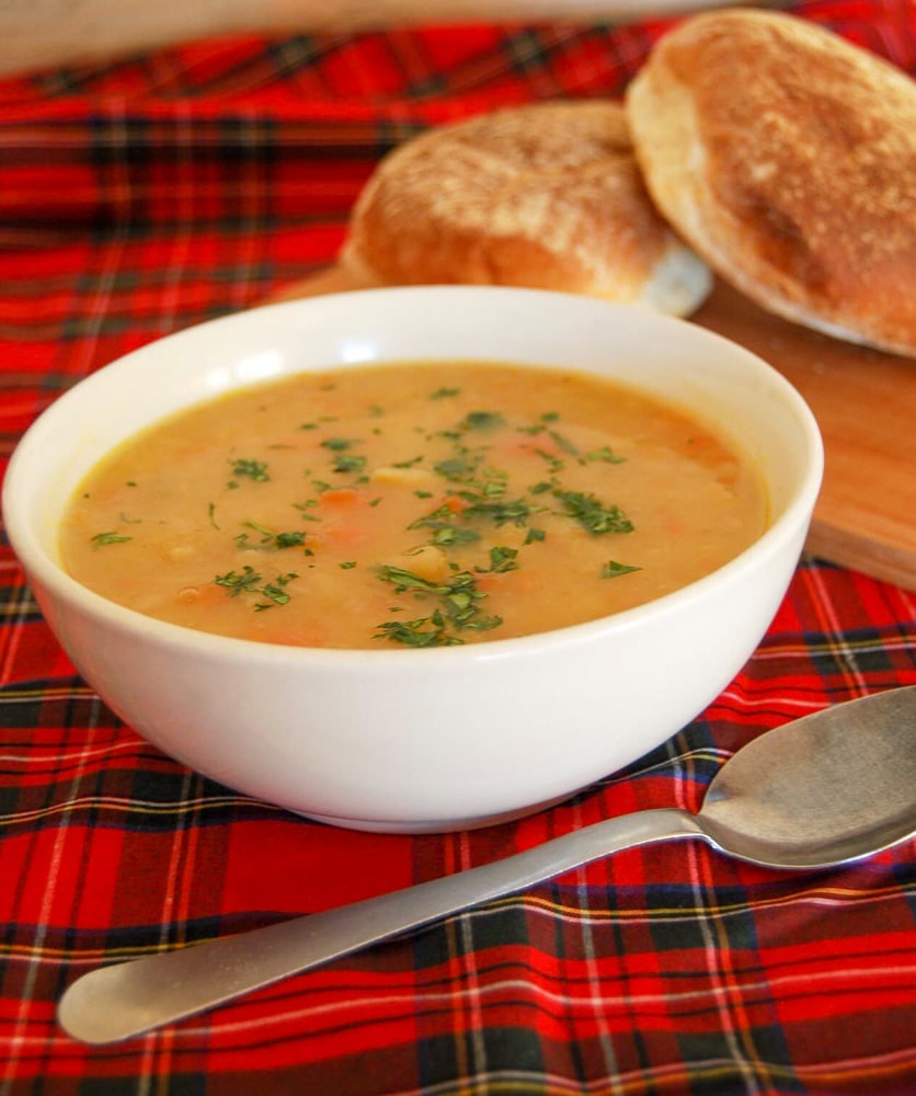 A bowl of tattie and carrot soup sprinkled with chopped parsley and two white rolls on a wooden serving board on a tartan tablecloth.