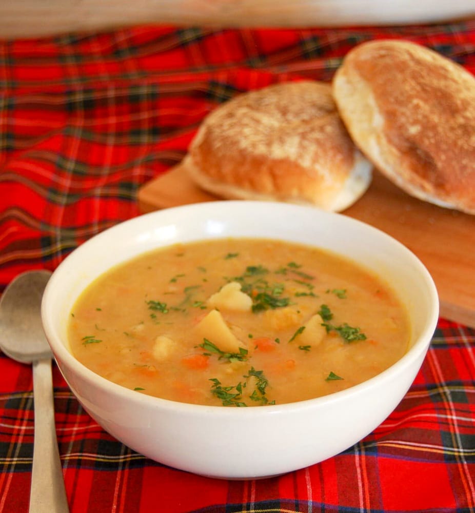 A white bowl of tattie soup sprinkled with chopped parsley with white rolls and a tartan tablecloth in the background.