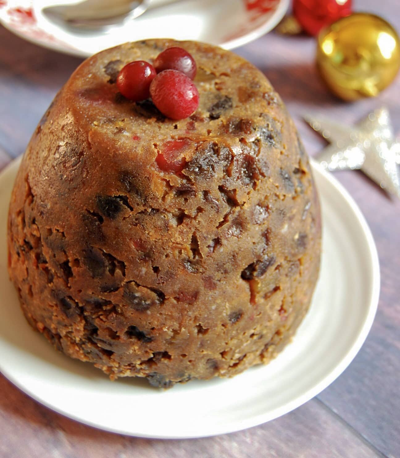 a Christmas pudding topped with fresh cranberries on a white plate, a silver star decoration and red and gold baubles.