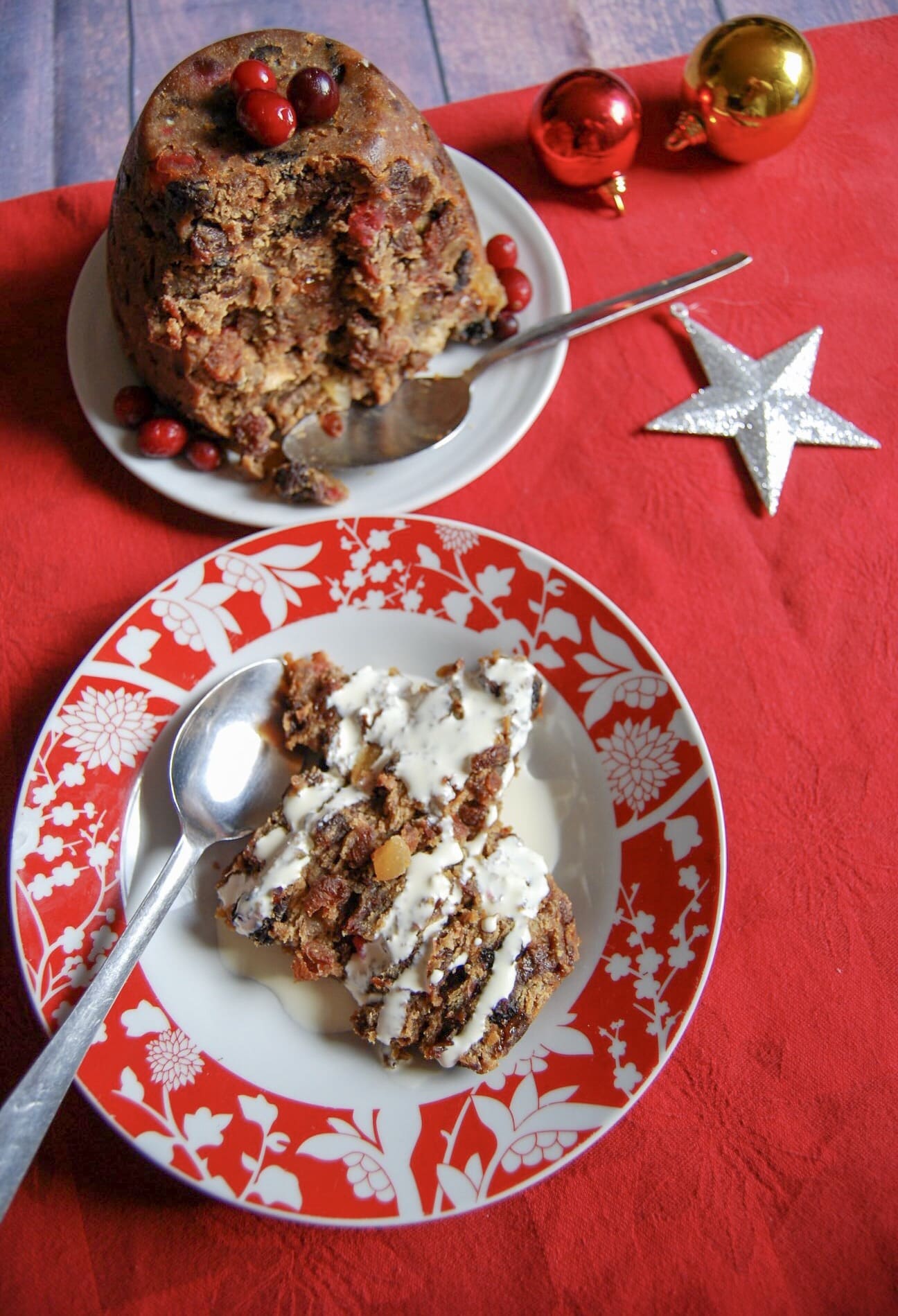 a flat lay picture of a Christmas pudding topped with cranberries on a white plate with a spoon, a red and white bowl with a slice of Christmas pudding and cream and red and gold baubles and a silver star on a red tablecloth.