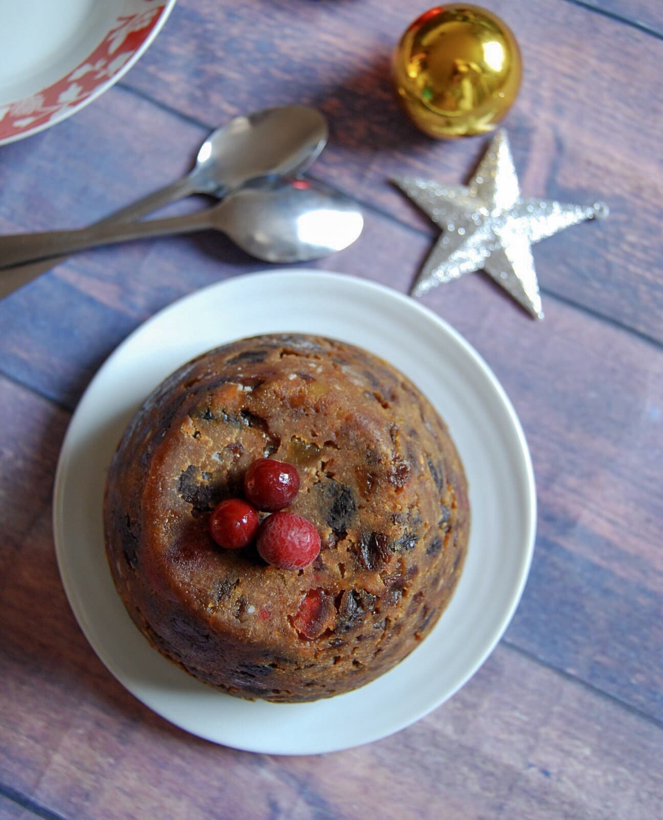 an overhead picture of a Christmas pudding topped with fresh cranberries on a white plate, two silver spoons, a silver star and gold bauble and the corner of a red and white bowl.