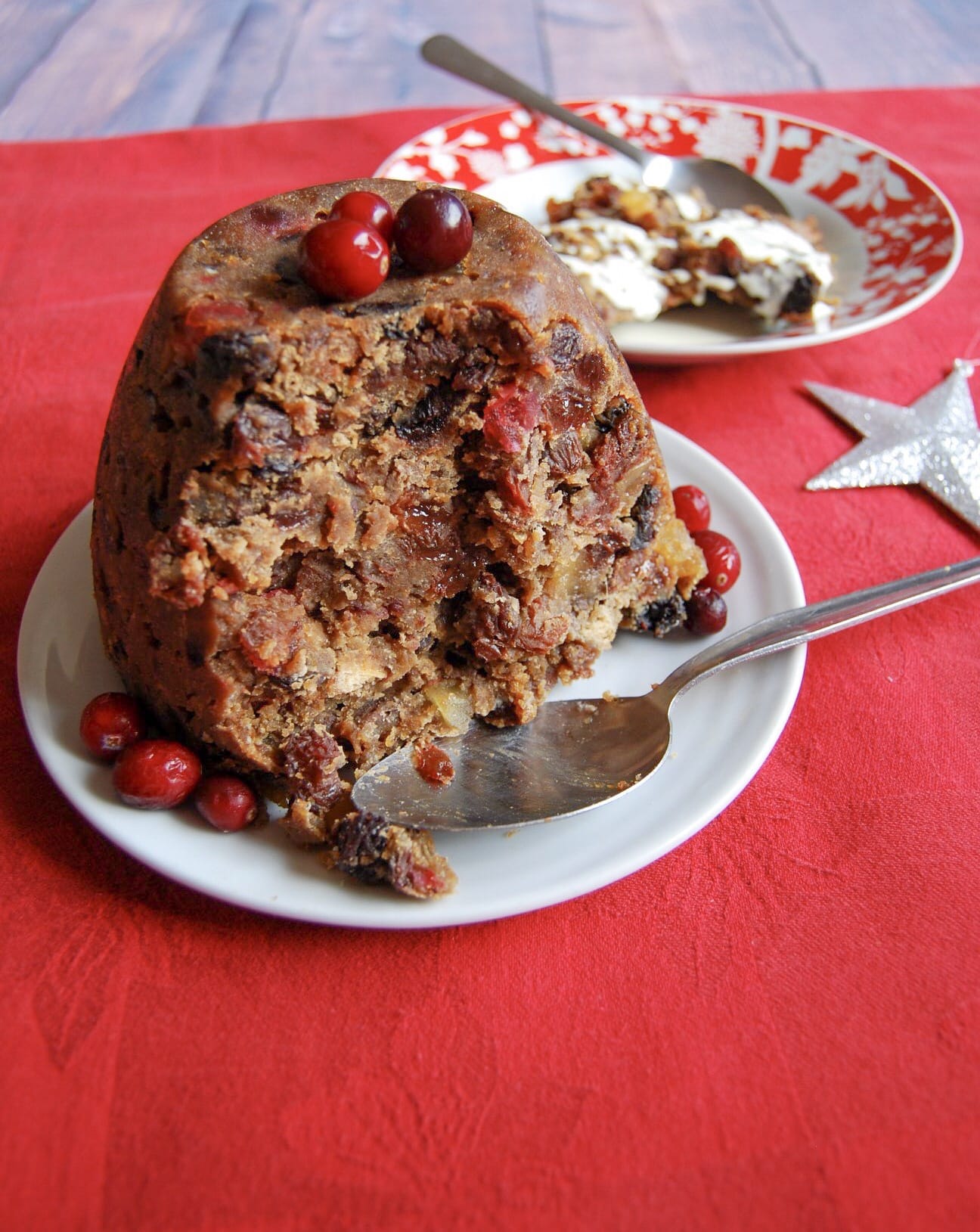 a Christmas pudding topped with fresh cranberries on a white plate with a serving spoon on a red tablecloth.  A red and white bowl with a slice of Christmas pudding and cream with a spoon can be seen in the background.