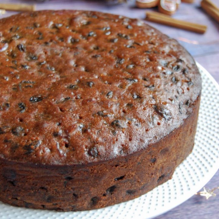 A round Amaretto fruit cake on a white plate. Christmas decorations and star lights can be seen in the background.