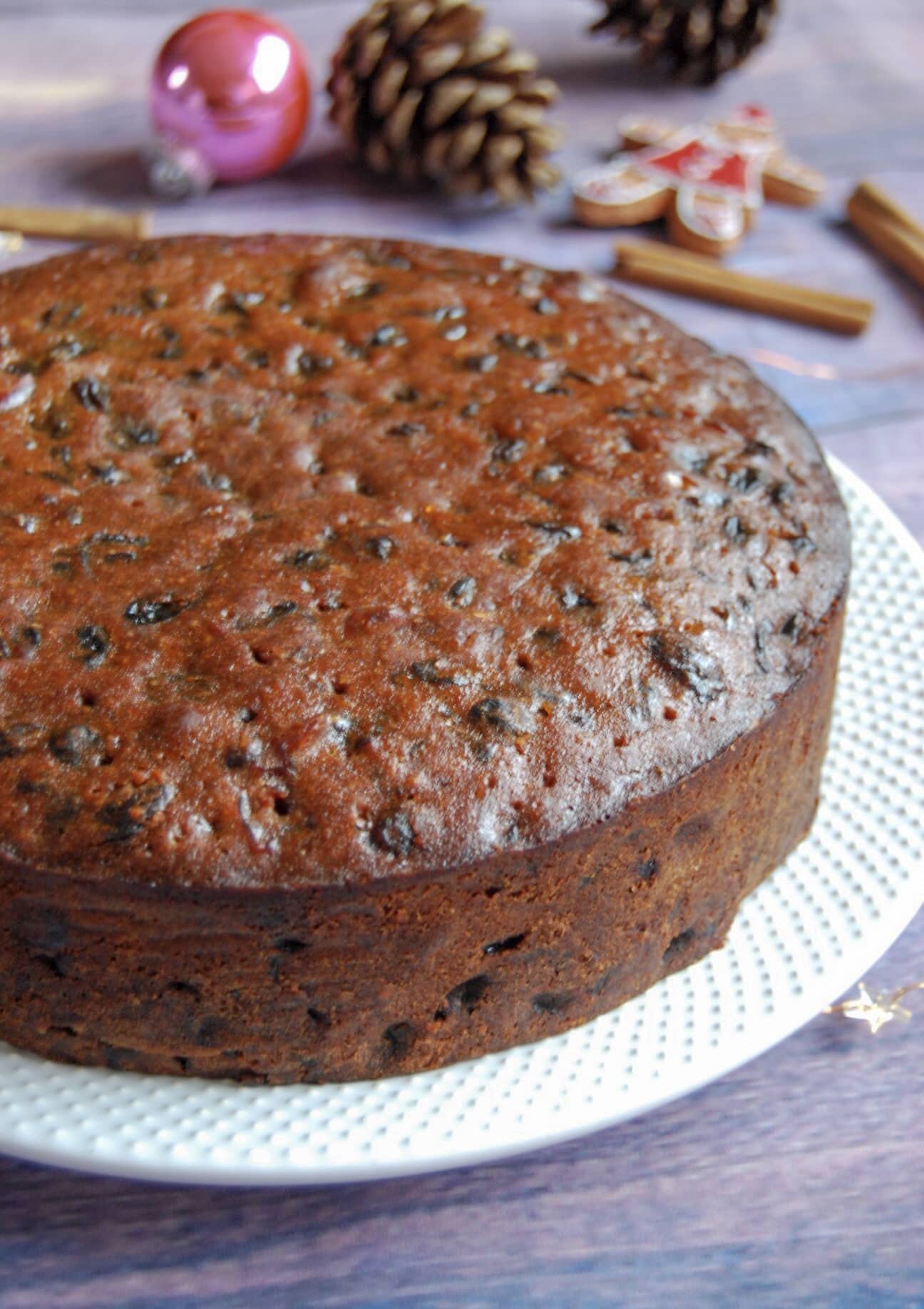 A round Amaretto fruit cake on a white plate. Christmas decorations and star lights can be seen in the background.