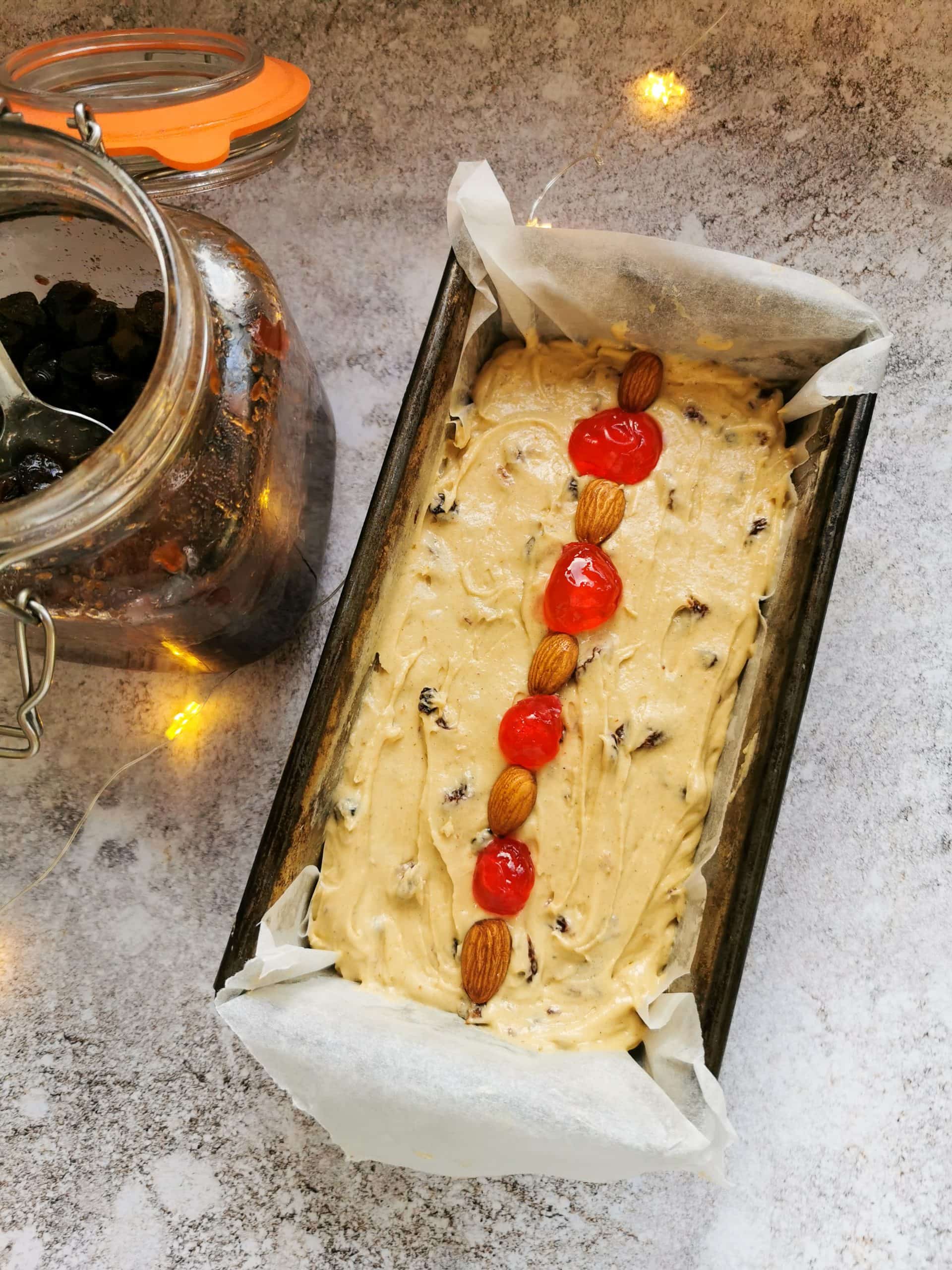 An unbaked fruit cake in a loaf tin studded with whole almonds and red glace cherries.