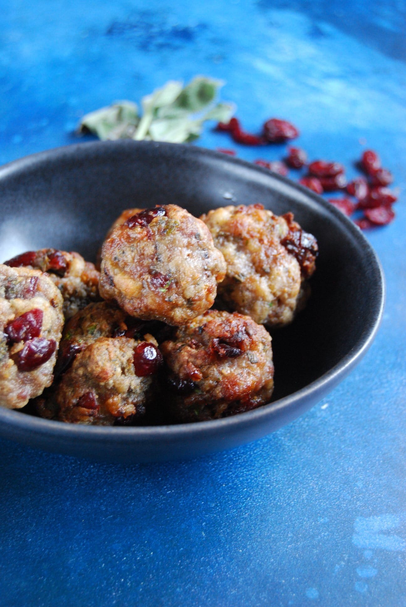 a small black bowl of cranberry studded sausagemeat stuffing balls on a dark blue background, a bunch of fresh sage and a handful of dried cranberries