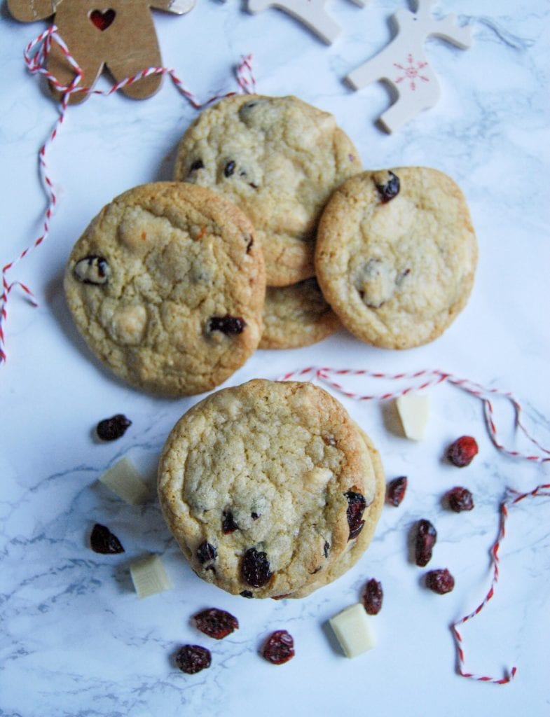 A flat lay photo of white chocolate cranberry cookies on a white marbled background. Dried cranberries and white chocolate is scattered around the cookies for decorative effect.