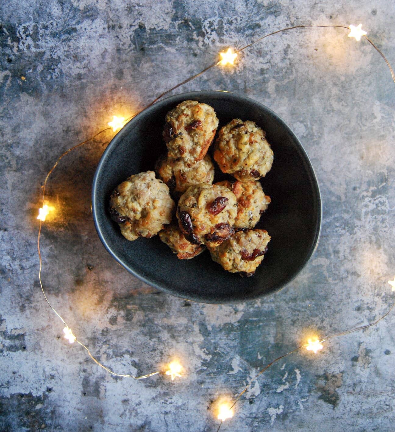 a black bowl filled with sausage and sage stuffing balls studded with cranberries surrounded by gold fairy lights on a grey background.