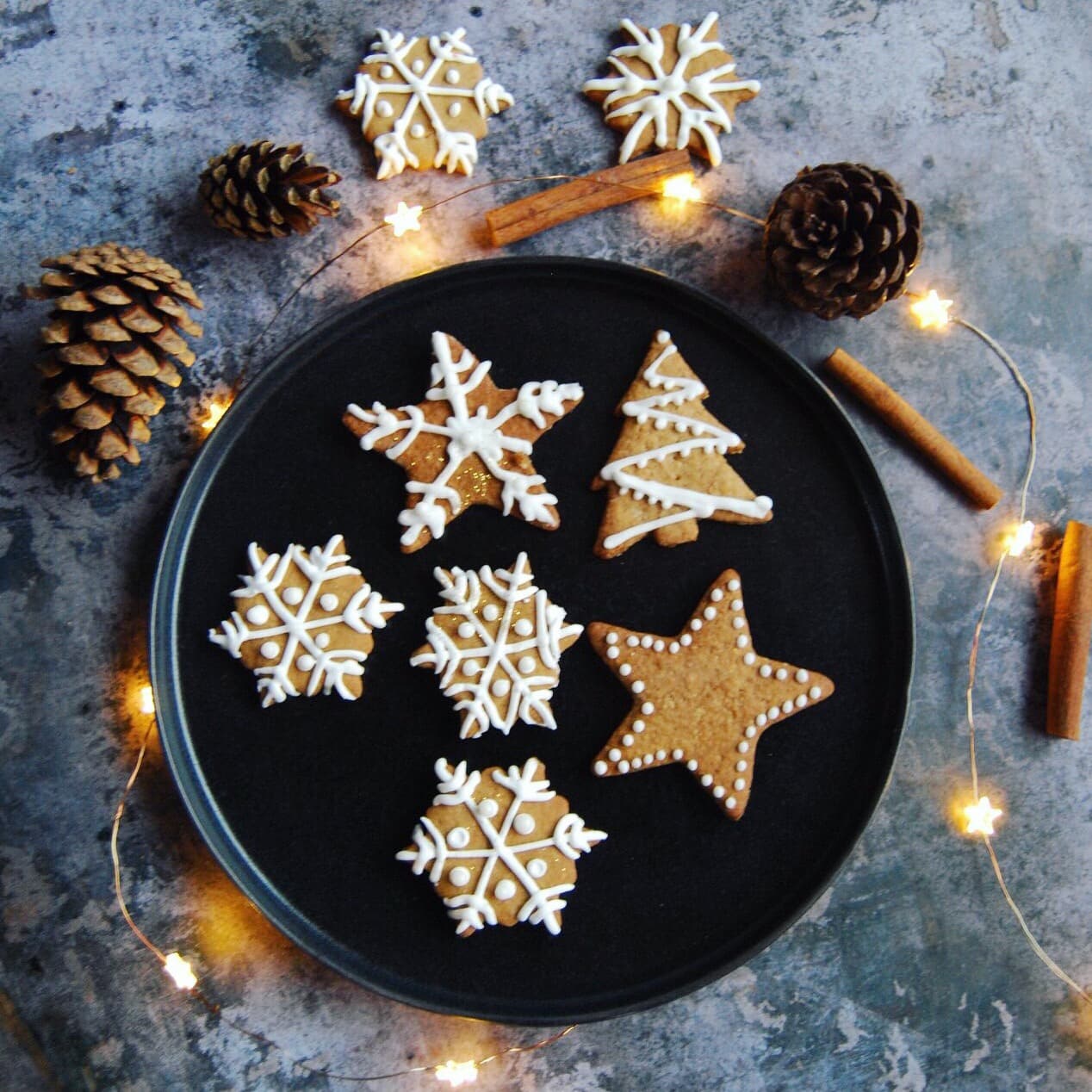 A photo of easy Iced Gingerbread Biscuits on a black plate and grey background with fairy lights, pine cones and cinnamon sticks