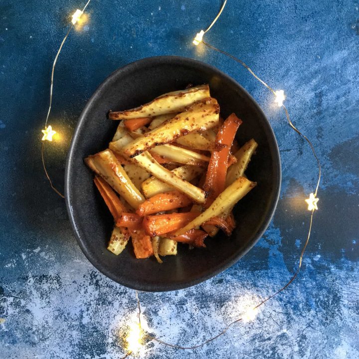 a bowl of honey mustard roasted parsnips and carrots on a blue background with Christmas fairy lights