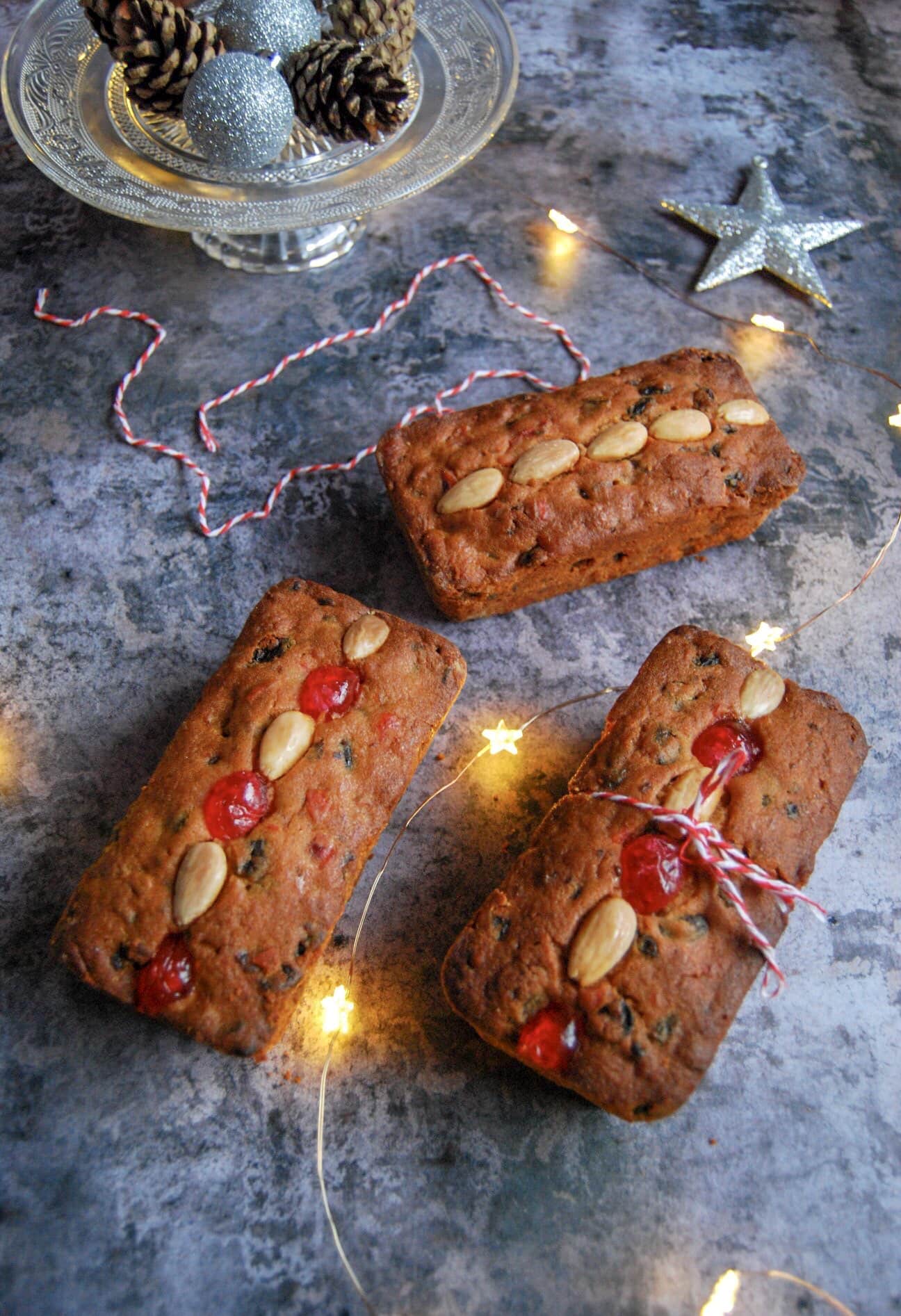 Three mini mincemeat loaf cakes studded with blanched almonds and cherries on a grey background with fairy lights and Christmas decorations