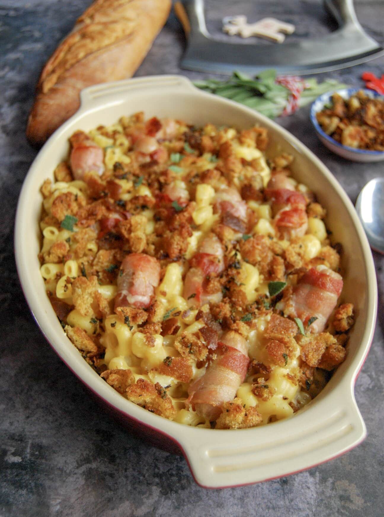 an oval dish of macaroni cheese with bacon wrapped sausages and topped with crispy sage breadcrumbs.  A French stick of bread can be seen in the background with a bunch of fresh sage