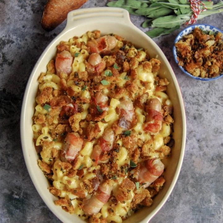 A flat lay photo of a gratin dish of pigs in blankets macaroni cheese - mac and cheese with bacon wrapped sausages and crispy, golden sage breadcrumbs