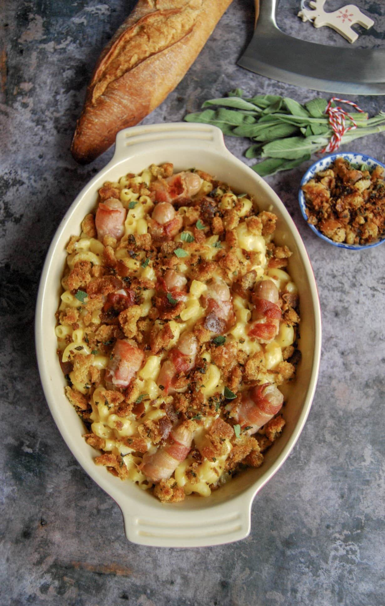 A flat lay photo of a gratin dish of pigs in blankets macaroni cheese - mac and cheese with bacon wrapped sausages and crispy, golden sage breadcrumbs