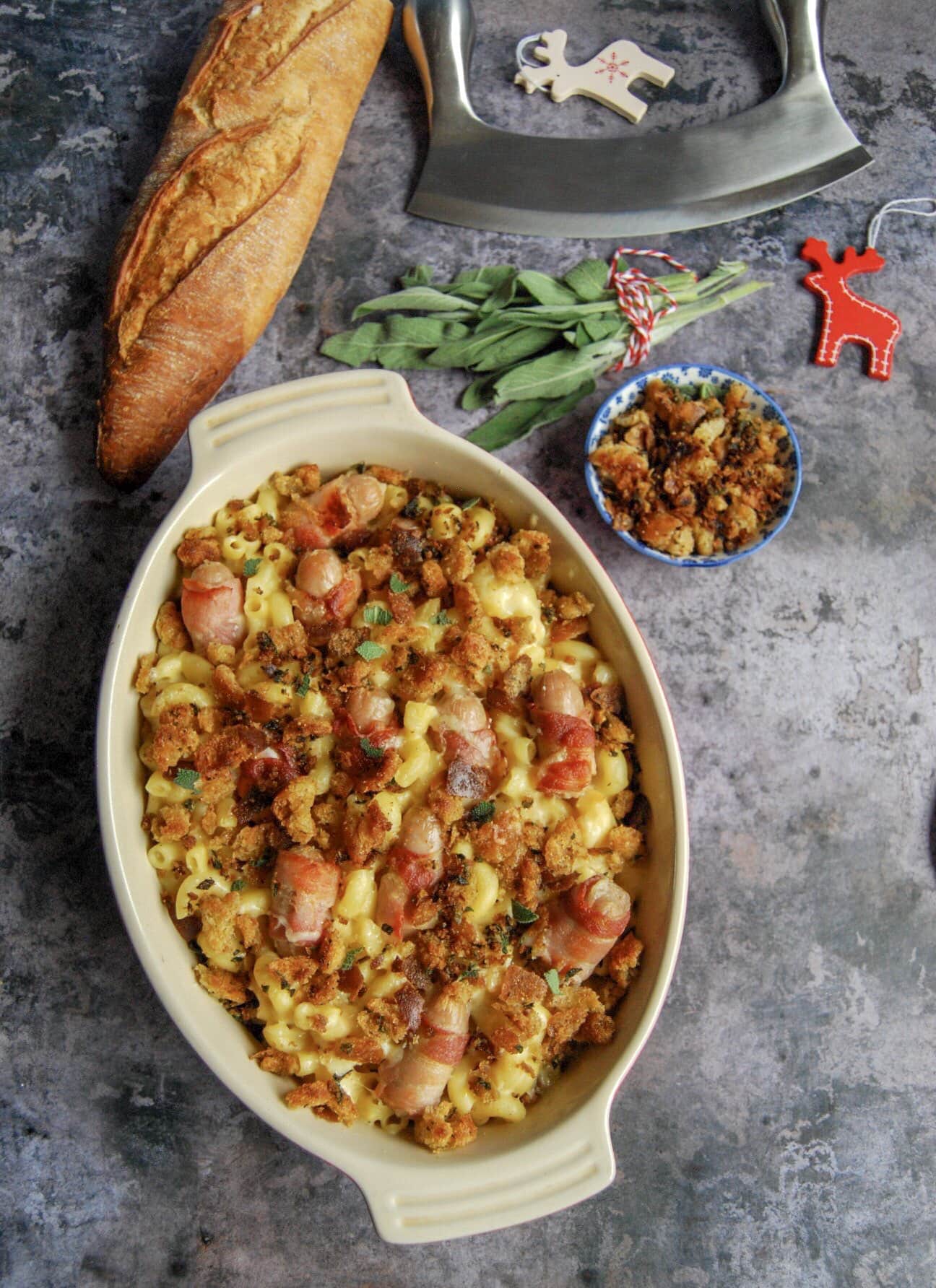 An oval casserole dish of macaroni cheese with bacon wrapped sausages and crispy sage breadcrumbs on a grey background. A French bread stick and a bunch of sage can be seen at the top of the picture