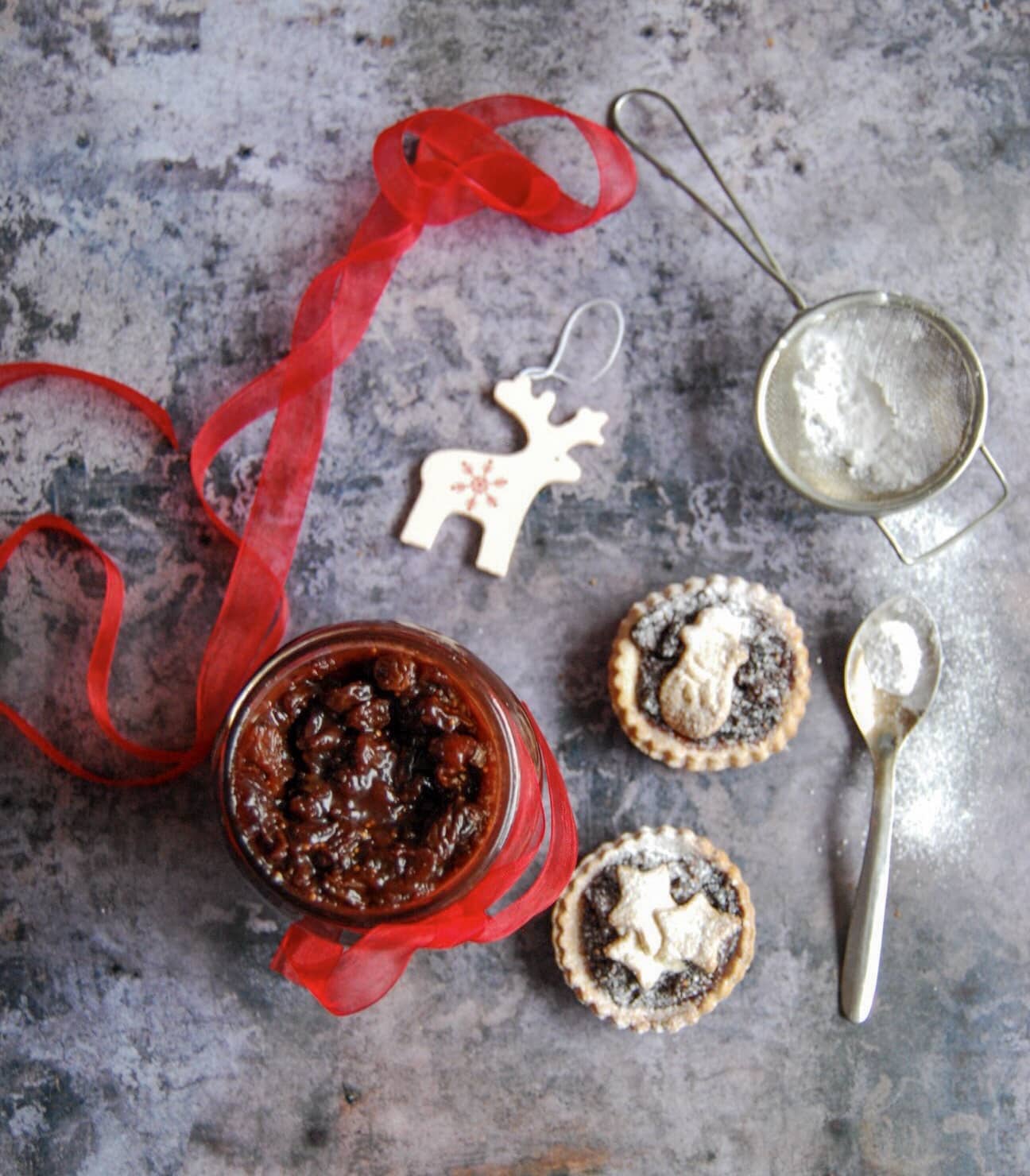 A jar of mincemeat with a red ribbon tied around the jar. Mince pies dusted with icing sugar are beside the jar. 