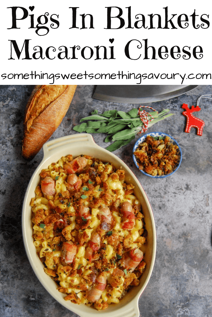 A pinterest pin with the words "pigs in blankets macaroni cheese" with a picture of an oval gratin dish filled with mac and cheese with bacon wrapped sausages, a French bread stick, a bunch of fresh sage and a small bowl of croutons.