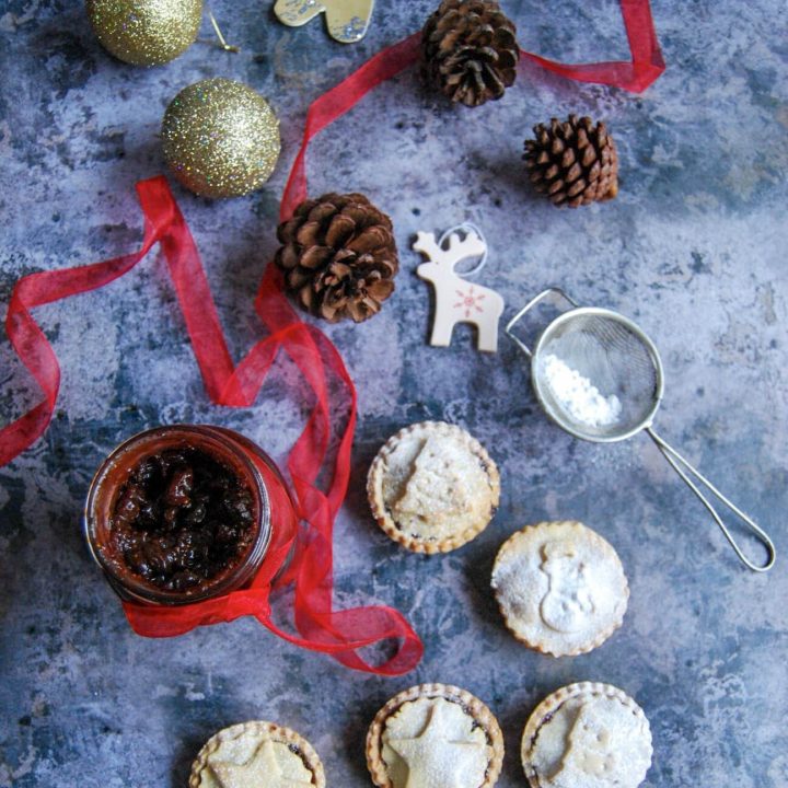 A jar of chocolate and cherry mincemeat and mince pies dusted with icing sugar. Christmas baubles and decorations are scattered around in a decorative fashion