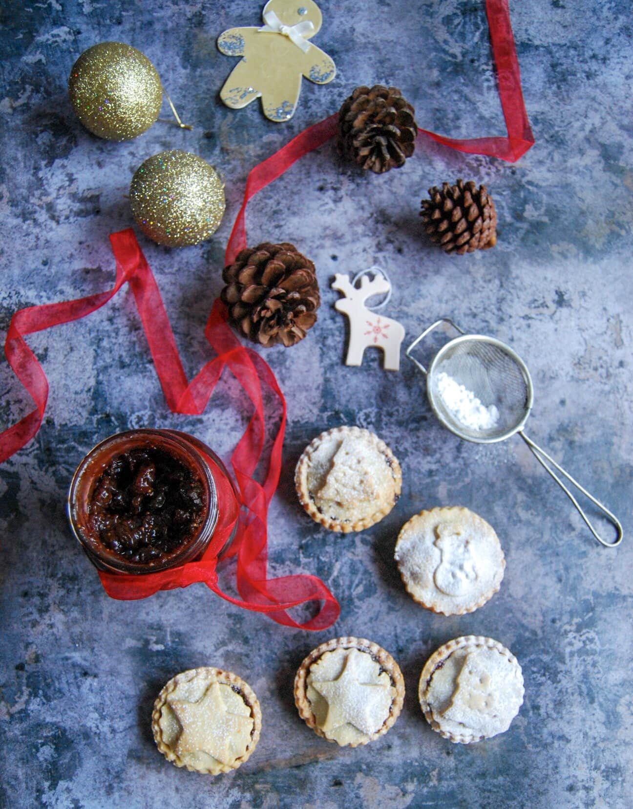 A jar of chocolate and cherry mincemeat and mince pies dusted with icing sugar. Christmas baubles and decorations are scattered around in a decorative fashion