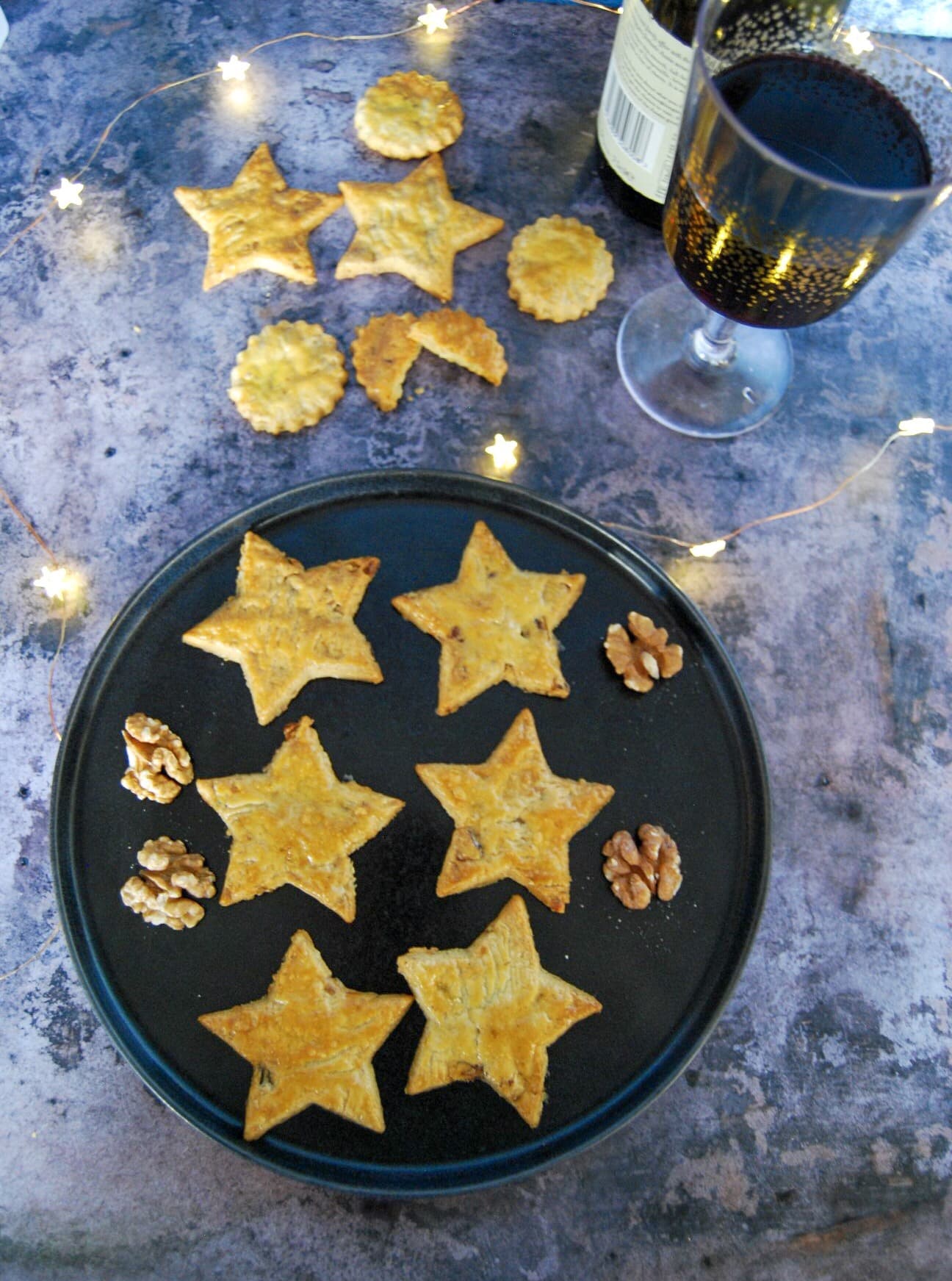 A black plate of cheese star biscuits, walnuts, a glass of red wine and golden fairy lights