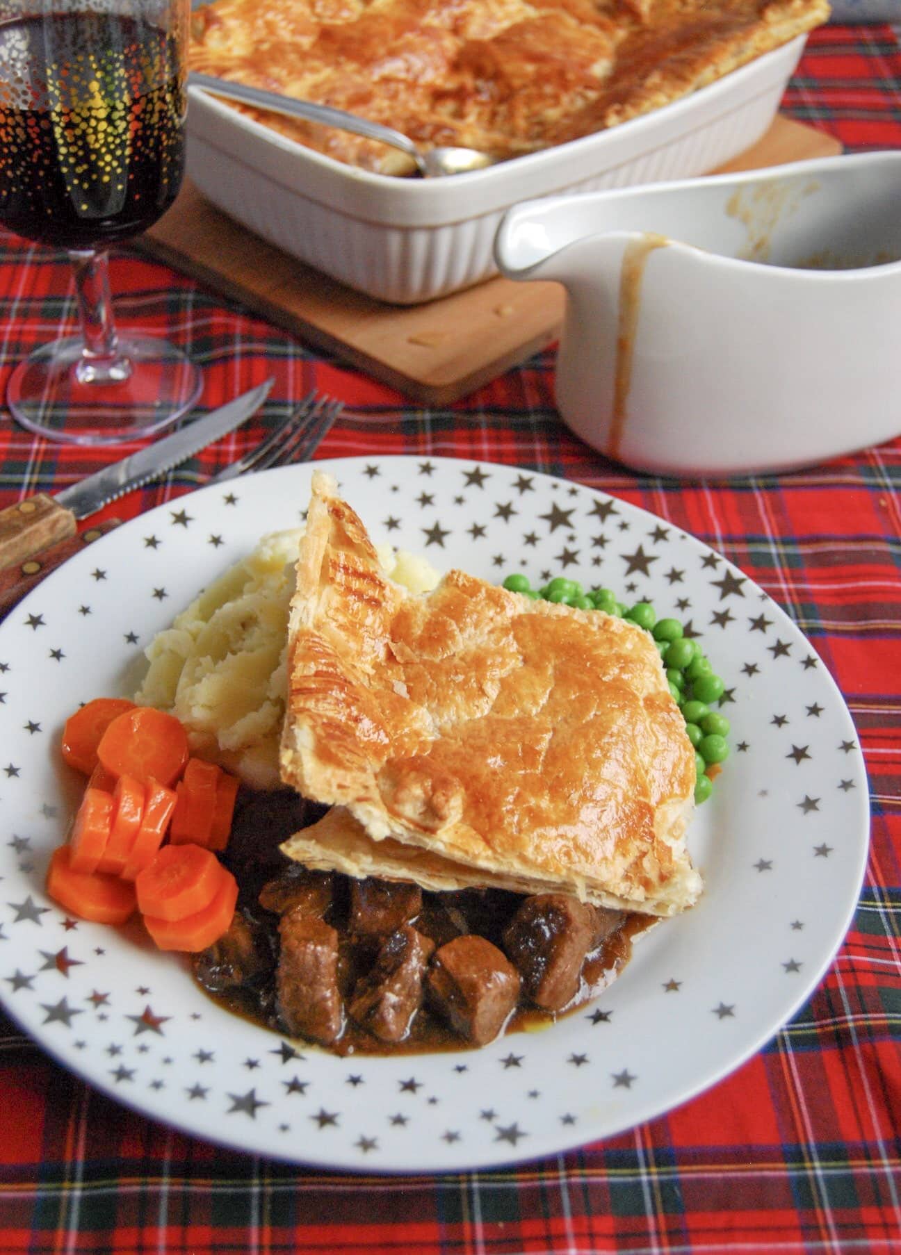 Steak pie with a puff pastry lid and chunks of beef in a rich gravy on a gold and white star place with mashed potatoes, carrots and peas. 