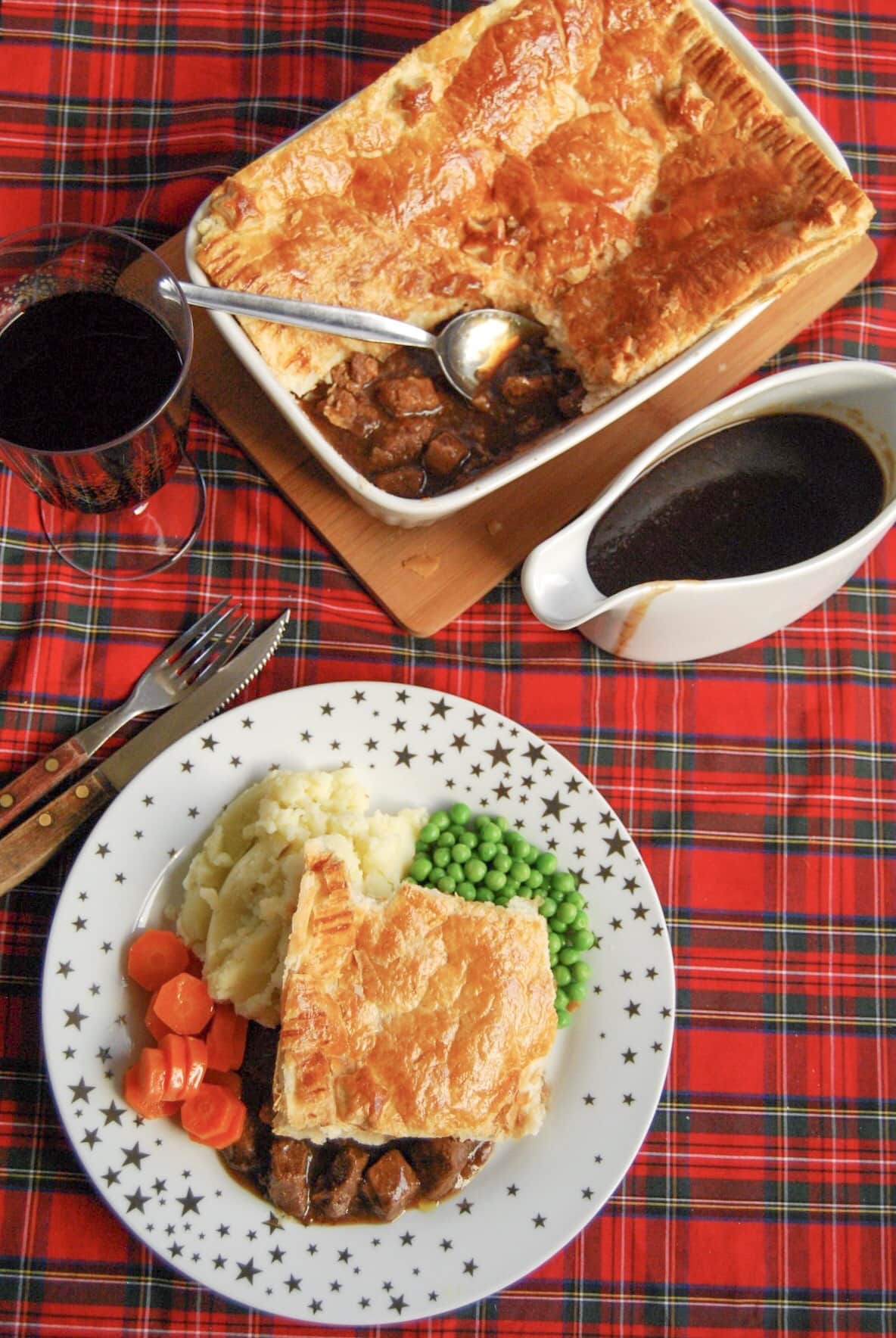 A flatlay photo of a gold and white star plate of steak pie with a puff pastry lid and beef chunks in gravy with mashed potatoes, peas and carrots. A jug of gravy and a large steak pie can also be seen in the top of the photo.