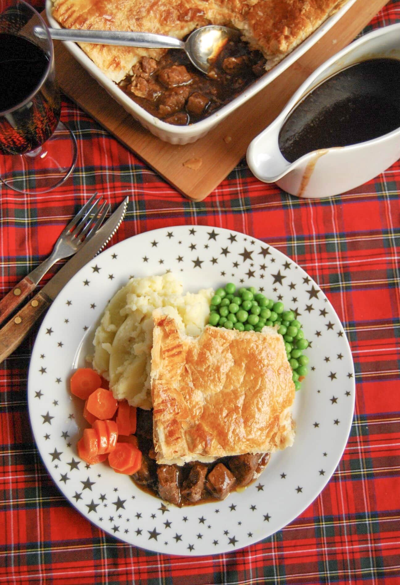 A flatlay photo of a gold and white star plate of steak pie with a puff pastry lid and beef chunks in gravy with mashed potatoes, peas and carrots. A jug of gravy and a large steak pie can also be seen in the top of the photo.