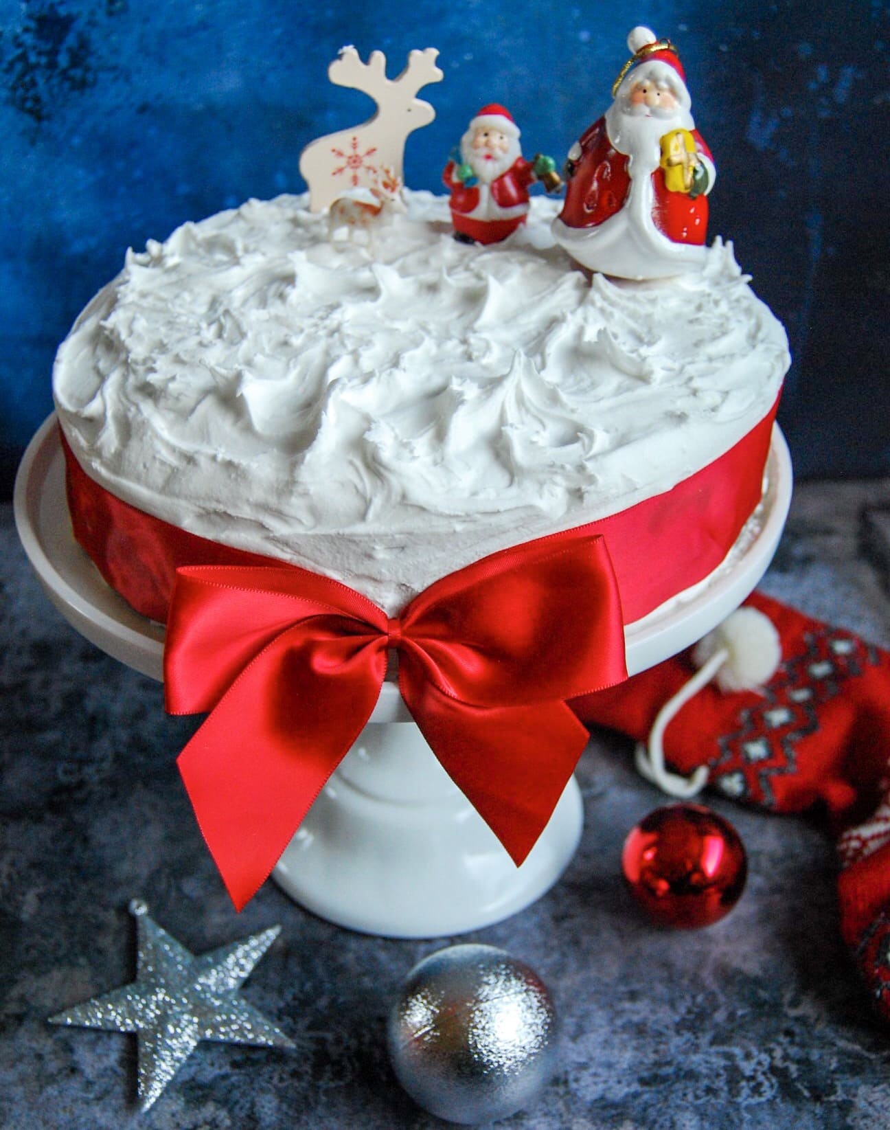 A Christmas cake decorated with white royal icing on a white cake stand. The cake has a prety red bow tied around it and it topped with Santa and Reindeer figures