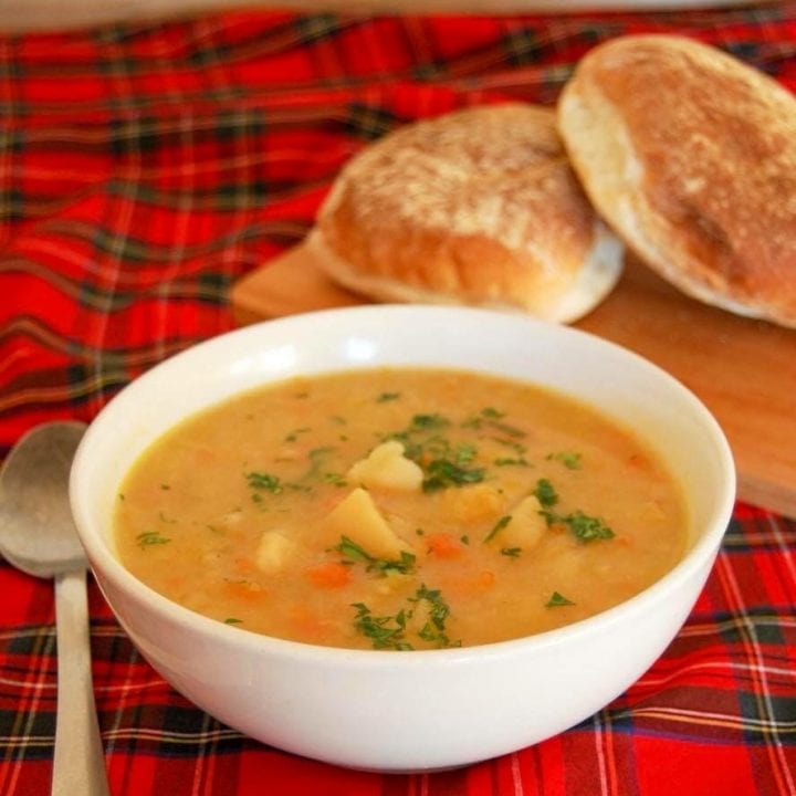A white bowl filled with tattie soup on a tartan tablecloth with bread rolls on the side.