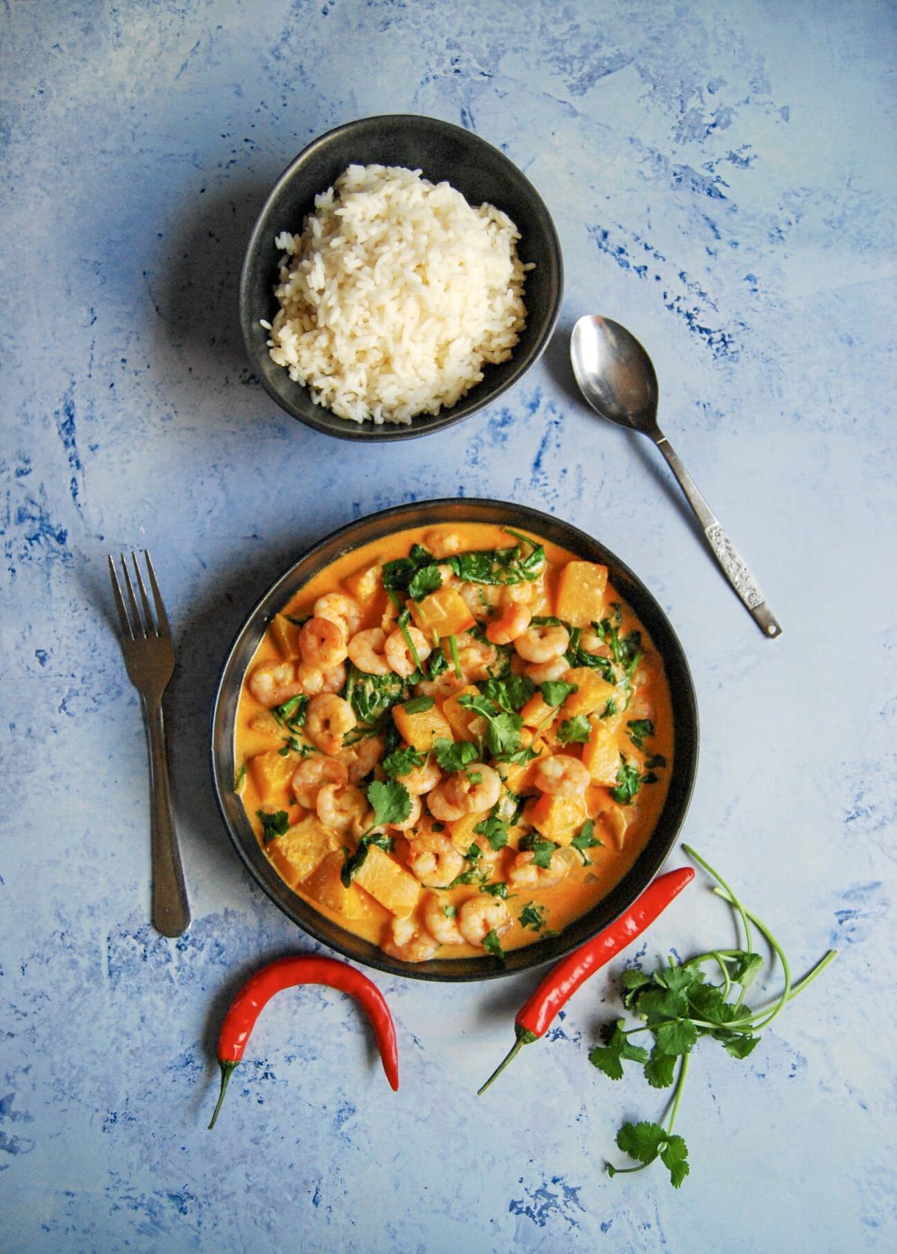 A flay lay photo - blue background with a bowl of prawn red Thai curry sprinkled with fresh chopped coriander. 