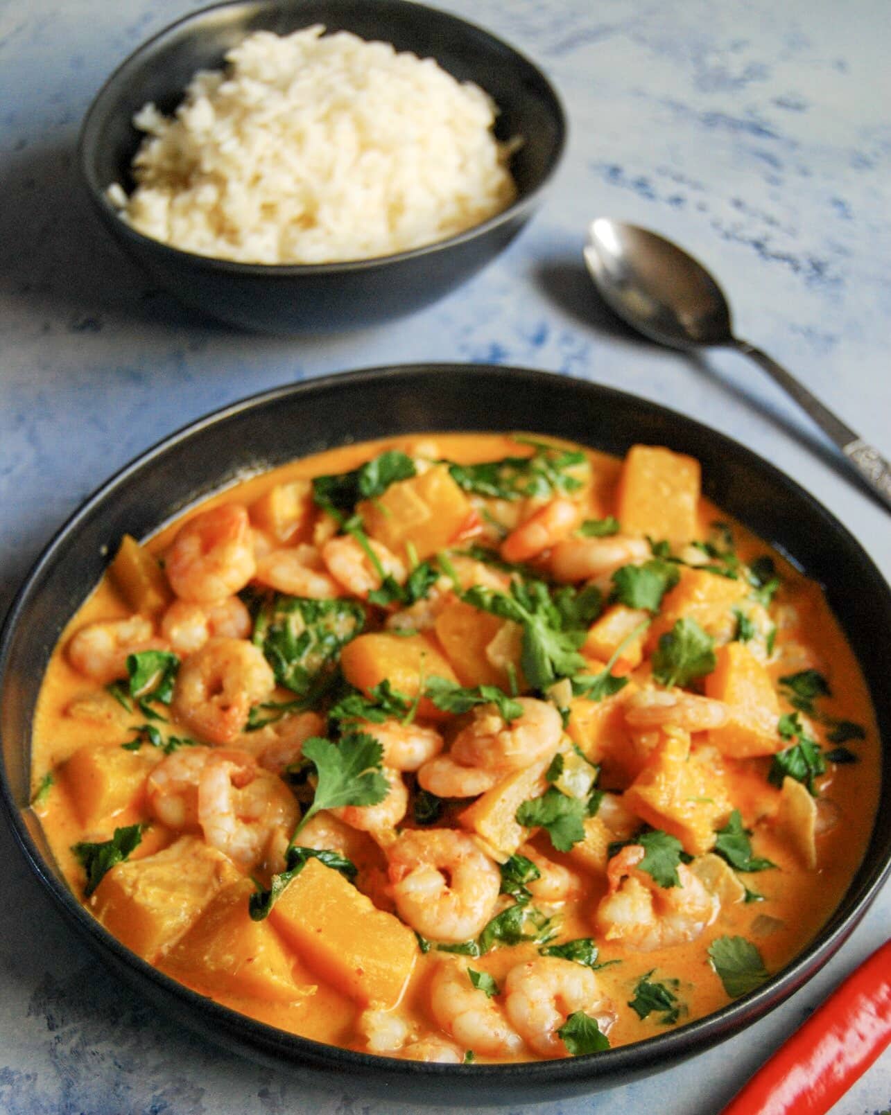 A bowl of Thai red curry with prawns, pumpkin, spinach sprinkled with chopped coriander with a bowl of rice in the background. 