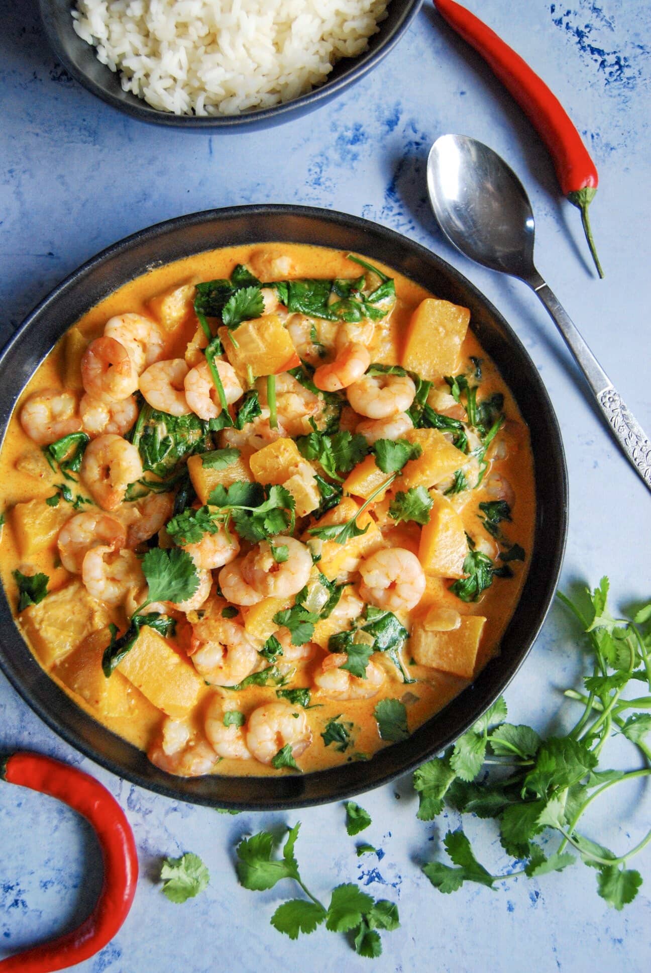 A flat lay photo - blue background with red chillies, chopped coriander and a bowl of prawn curry sprinkled with chopped coriander