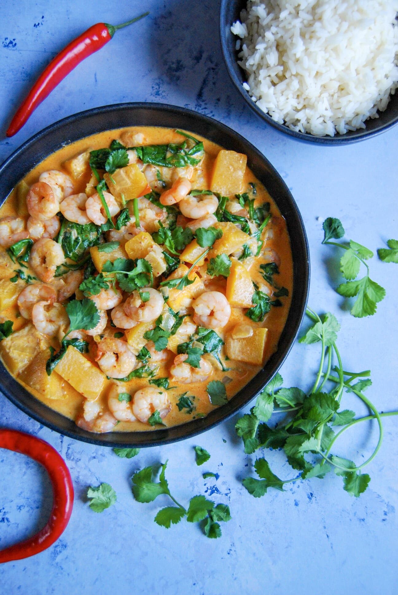 A flay lay photo - blue background with a bowl of prawn red Thai curry sprinkled with fresh chopped coriander. A bowl of rice can be seen in the top right hand corner and red chillies and coriander sprigs are placed around the bowls.