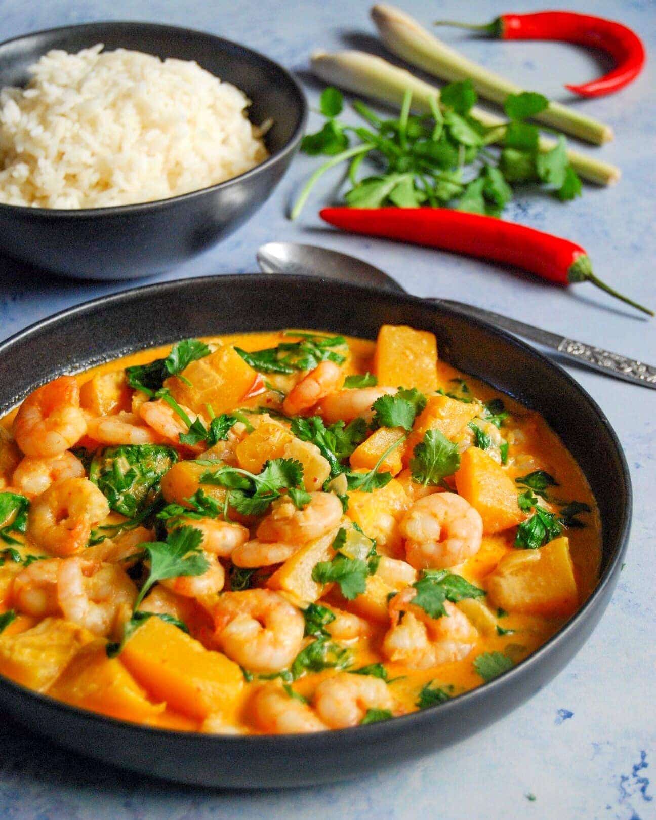 A bowl of Thai red curry with prawns, pumpkin, spinach sprinkled with chopped coriander with a bowl of rice in the background. 