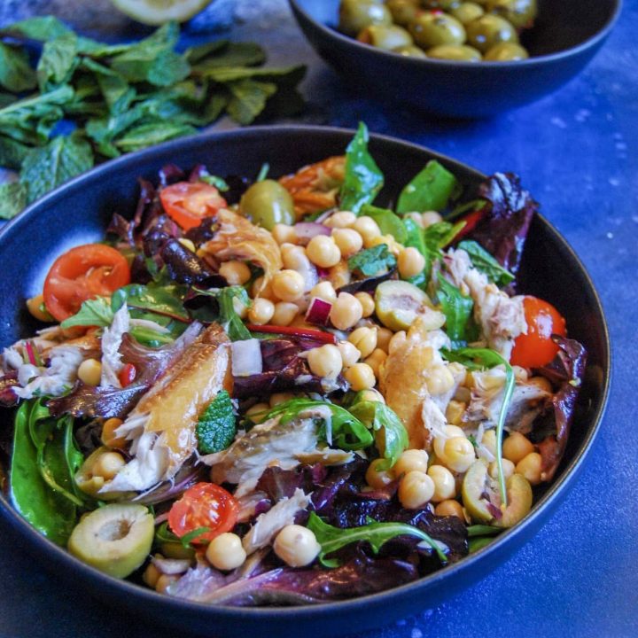 A bowl of mackerel and chickpea salad on a blue background. A bowl of green olives and fresh herbs are seen in the background.