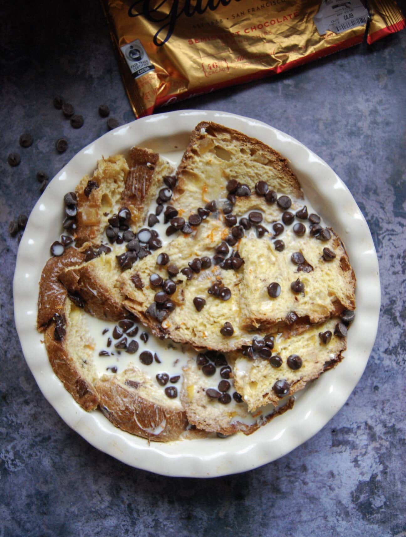 A round dish with panettone slices soaked in egg custard and sprinkled with orange zest and chocolate chips. A packet of chocolate chips are sitting above the pie dish