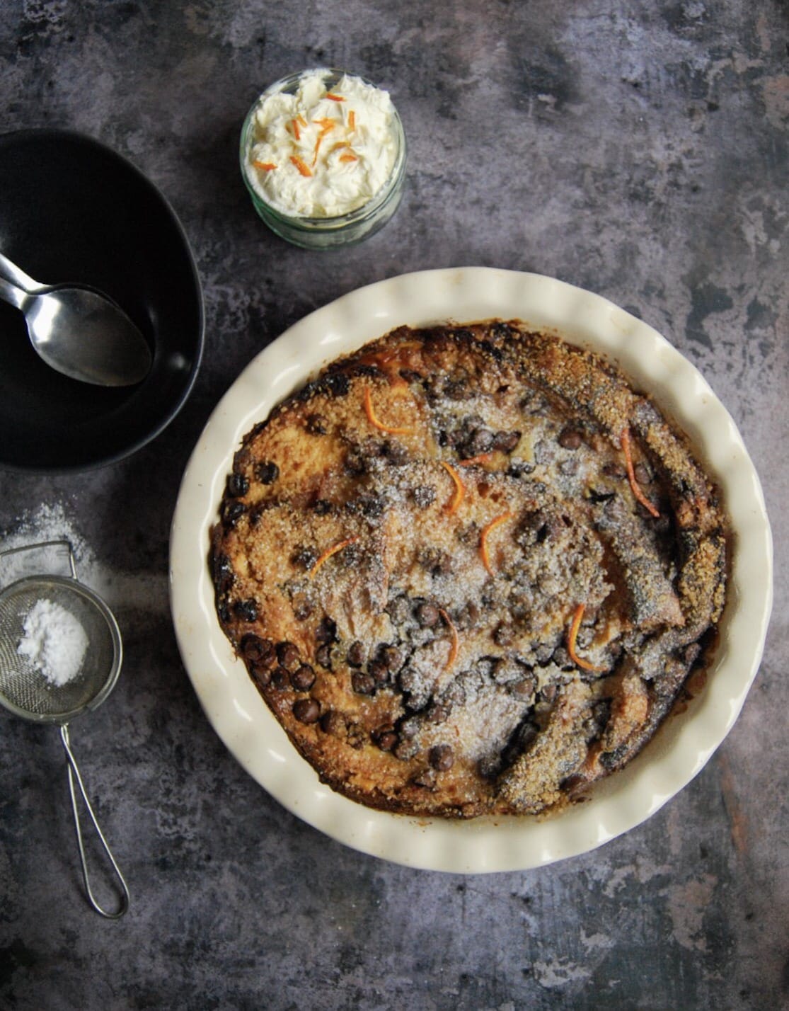a panettone bread pudding in a round white dish, a black bowl with spoons and a little pot of whipped cream topped with orange zest