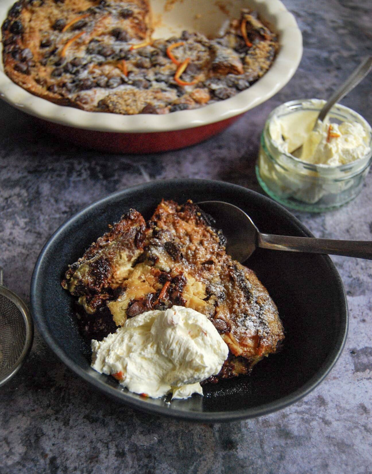 chocolate orange bread pudding with chocolate chips and whipped cream in a small black round bowl. A round dish of the pudding and a small dish of whipped cream