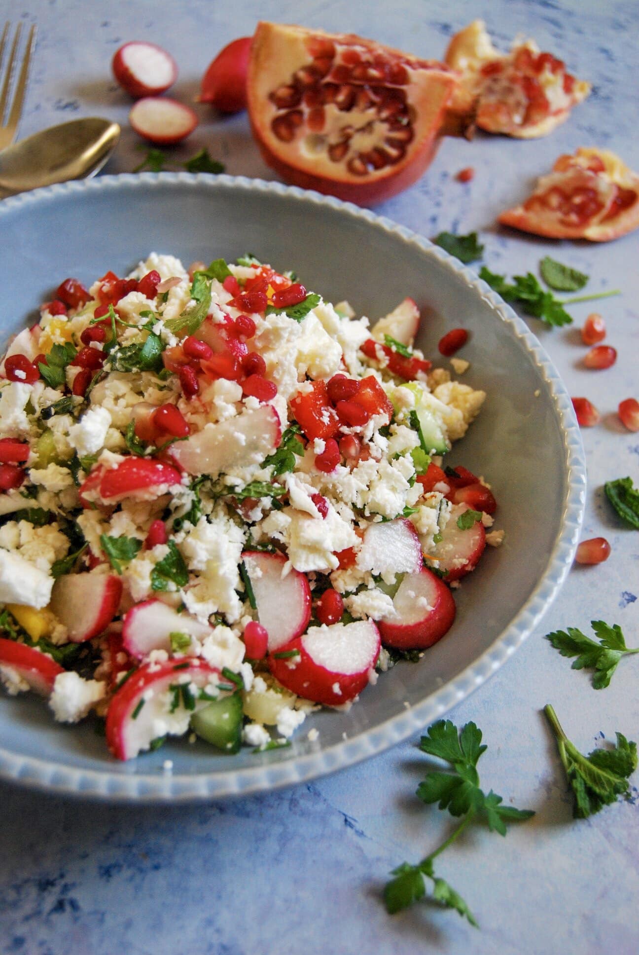 A bowl of cauliflower cous cous salad with radishes, pomegranate seeds and green herbs on a light blue background.