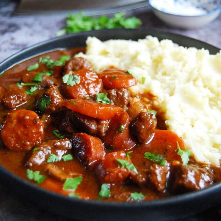 A plate of slow cooker beef and chorizo casserole with carrots and mashed potato on a grey background