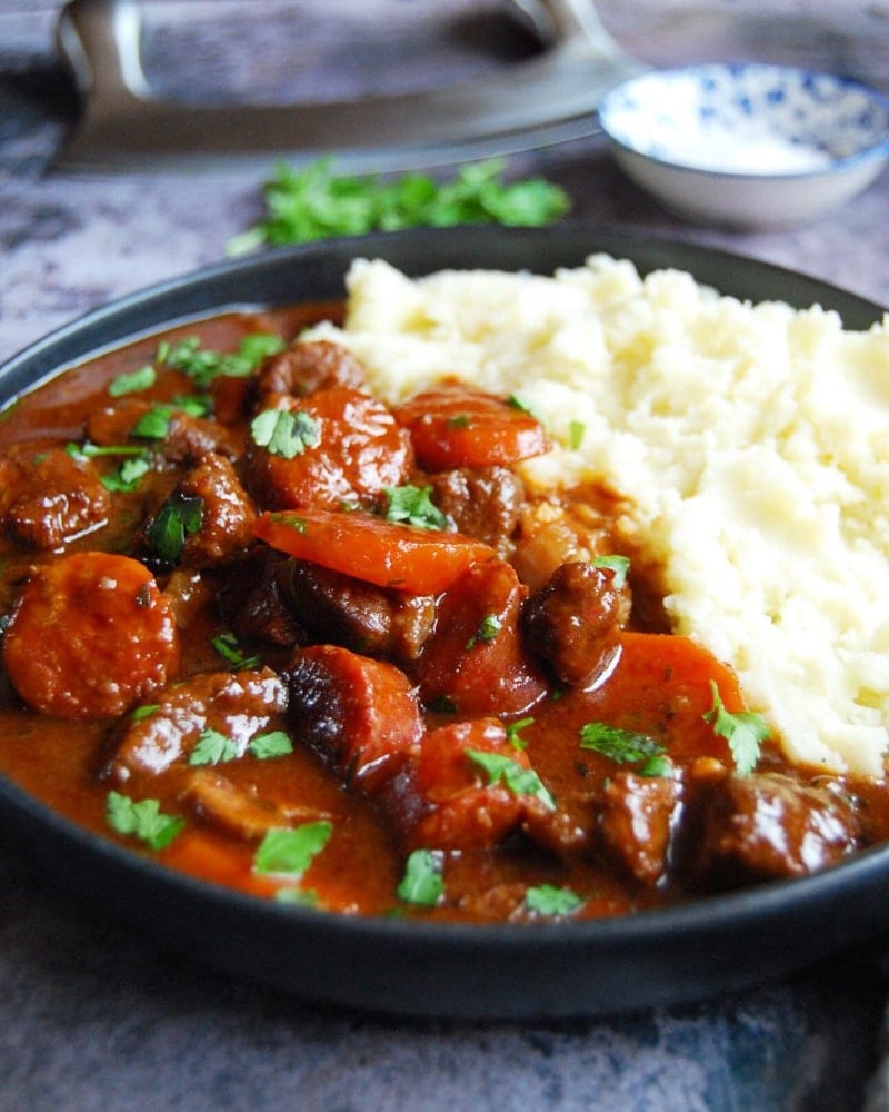 A plate of slow cooker beef and chorizo casserole with carrots and mashed potato on a grey background