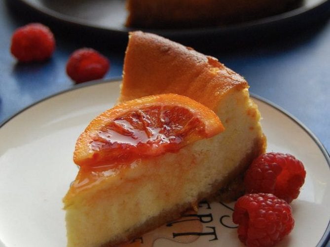 A slice of baked cheesecake decorated with a candied blood orange slice with fresh raspberries on a black and white plate